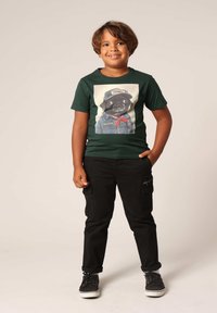 Groene katoenen t-shirt met een afbeelding van een hond in een pet en een jas; gecombineerd met zwarte cargobroek en zwarte sneakers.