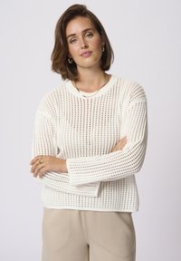 Pull en tricot blanc avec un motif en open weave, col rond et manches longues. Porté avec un bas beige. Texture lisse, coupe décontractée.