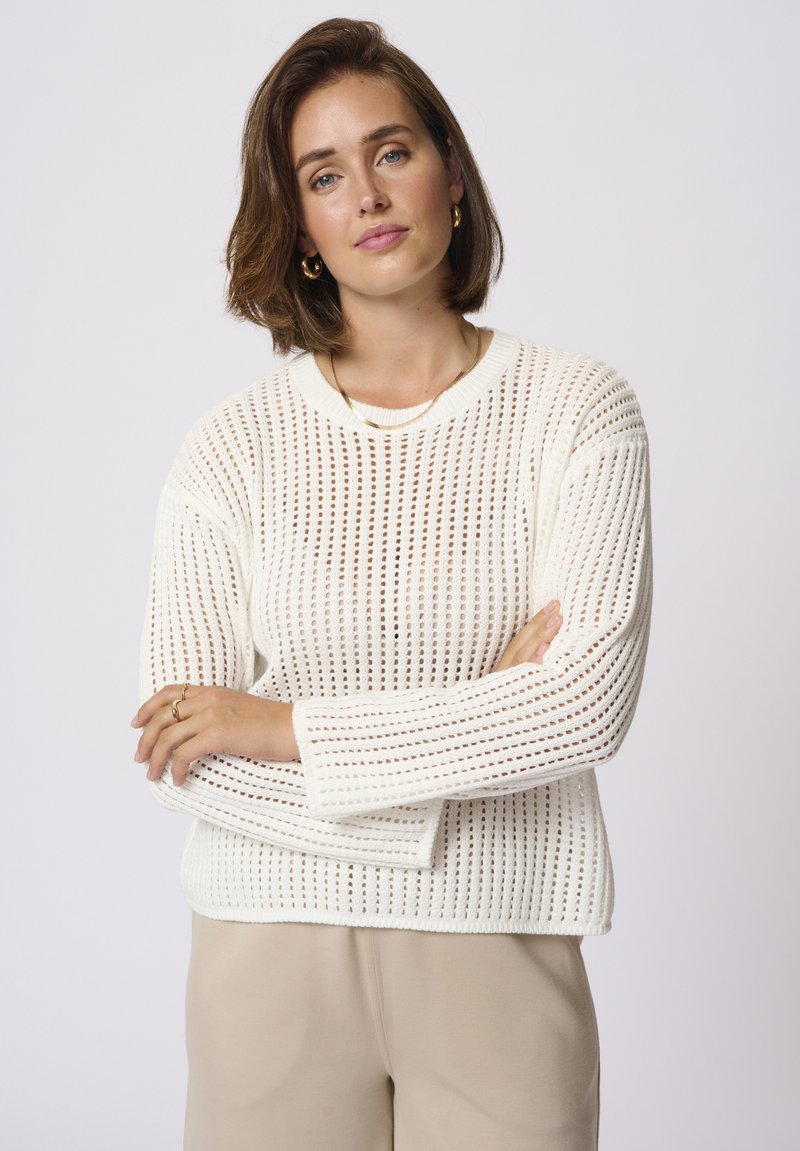 Pull en tricot blanc avec un motif en open weave, col rond et manches longues. Porté avec un bas beige. Texture lisse, coupe décontractée.