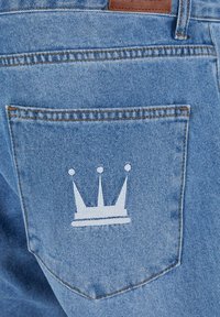 Hellblaue Denim-Jeans haben eine Gesäßtasche mit einem weißen Kronenmotiv. Der Stoff hat eine klassische Textur mit gelben Nähakzenten.