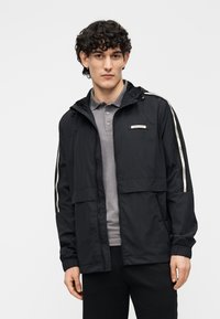 Chaqueta negra ligera con capucha, cremallera frontal y dos bolsillos laterales; presenta rayas blancas verticales a lo largo de las mangas.