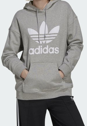 Osoba ubrana w szarą bluzę z kapturem Adidas z dużym białym logo typu trefoil oraz czarne spodnie Adidas, ręce w przedniej kieszeni, widoczna górna część ciała.