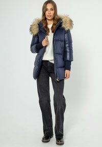 Marineblauwe pufferjack met een capuchon afgewerkt met bont, glanzend materiaal, ritsluiting en geribde manchetten, gedragen over een witte trui en donkere jeans.