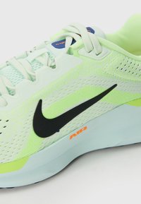 Svijetlozelena atletska tenisica s mrežastim gornjim dijelom, crni Nike logo, teksturirana potplat i narančasti akcent "AIR". Fleksibilan dizajn za udobnost i performanse.