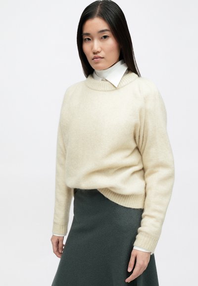 Κρέμα χρώματος, μαλακό πλεκτό πουλόβερ με στρογγυλή λαιμόκοψη και ριμπ μανίκια, συνδυασμένο με μια σκούρα πράσινη εφαρμοστή φούστα.