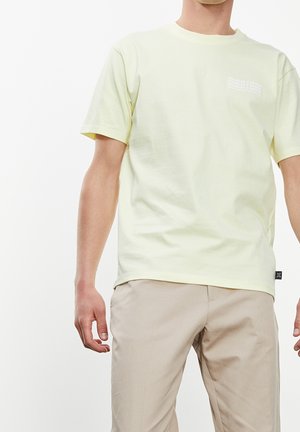 Hombre con camiseta de manga corta de color amarillo claro y pantalones beige, de pie frente a un fondo blanco liso.