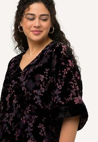 Musta samettipaita, jossa on kukkakuosi purppuran ja sinisen sävyissä, V-kaula-aukko ja lyhyet, käännetyt hihat. Pehmeä rakenne, laskeutuu sulavasti.