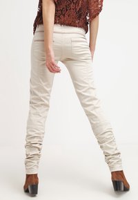 Beżowe spodnie slim-fit o gładkiej fakturze, z pionowymi szwami i marszczonym materiałem przy kostkach, w połączeniu z brązowymi botkami na obcasie.