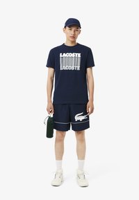 Camiseta navy con logo blanco de "LACOSTE", pantalones cortos navy con ribetes blancos y gráfico de cocodrilo, combinados con zapatillas blancas y una botella de agua verde.