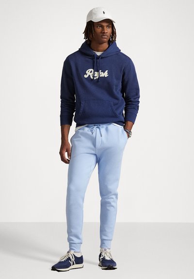Marineblå bomulds-hoodie med "Ralph" logo, lyseblå sweatpants, hvid cap og marineblå sneakers med creme-akcenter, stående mod en neutral baggrund.
