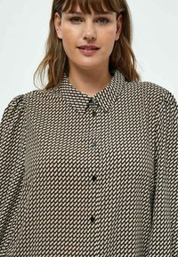 Button-up shirt in a geometric black, beige, and brown pattern; features a collar, black buttons, and puffed sleeves. Textured fabric.