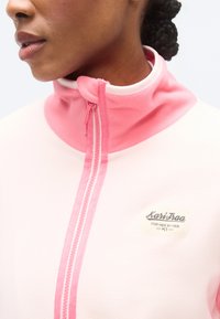 Ζακέτα από φλις με ροζ και κρεμ αποχρώσεις, ψηλό γιακά και ροζ φερμουάρ. Διαθέτει ετικέτα λογότυπου στο μπροστινό μέρος με κείμενο.