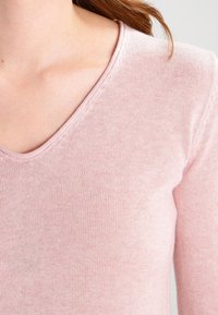 Różowy dziany top z długim rękawem i dekoltem w serek, o gładkiej fakturze i dopasowanym kroju, podkreślający miękkość tkaniny.