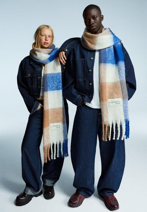 Mehrfarbiger Schaal aus Wollmischung mit blauen, weißen und sandfarbenen Streifen sowie Fransenenden. Getragen von Personen in Jeans-Outfits.