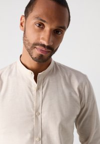 Jonge man met een korte baard en snor, gekleed in een beige overhemd met mandarin-kraag tegen een effen witte achtergrond.