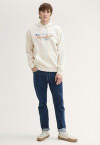 Felpa crema con tasca frontale, stampa blu e arancione, abbinata a jeans blu scuro e sneakers bianche con dettagli beige. Look casual.