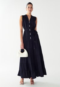 Maxi šaty v tmavomodrej farbe. Majú predné zapínanie na gombíky, viazanie v páse a textúrovanú vrstvenú látku. Skombinované s bielou kabelkou s oblým tvarom.