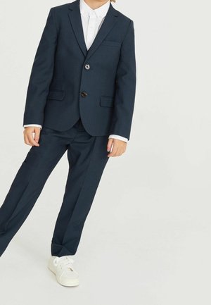 Bambino che indossa un abito blu scuro con camicia bianca e sneakers bianche, in piedi su uno sfondo chiaro con una gamba leggermente piegata.