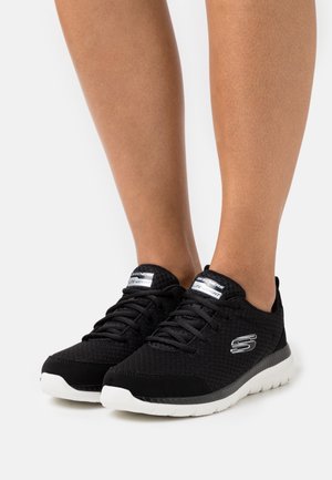 Chaussures de sport légères noires avec empeigne en mesh, panneaux en tissu texturé, semelle blanche, et logo argenté proéminent sur le côté.