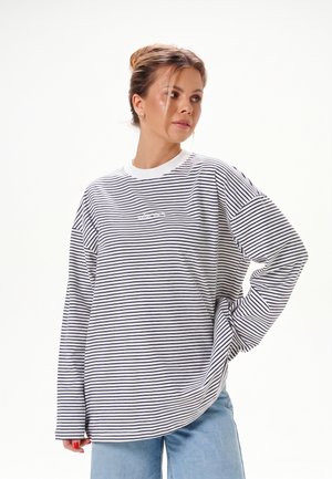 Femme avec les cheveux en chignon portant une chemise blanche oversize à rayures horizontales noires et un jean bleu clair, debout avec une main sur la hanche.