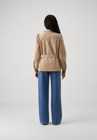En person med långt, vågigt hår bär en beige jacka med bälte och blå vida jeans, sedd bakifrån mot en neutral bakgrund.