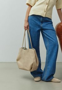 Geantă tote din piele bej cu o suprafață moale și texturată; două curele lungi; deschidere în partea de sus cu detalii fine de cusătură; asortată cu blugi albaștri cu croi larg.