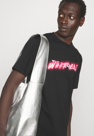 Fiatal férfi fekete pólóban, amelyen neon rózsaszín felirat „WHAT?” látható, és nagy fémes ezüst színű válltáskát visel, oldalra néz.