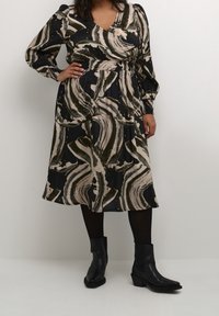 Robe à manches longues avec un décolleté en V profond, présentant un motif abstrait tourbillonnant en noir, beige et vert olive, cintrée à la taille. Bottines noires à la cheville.