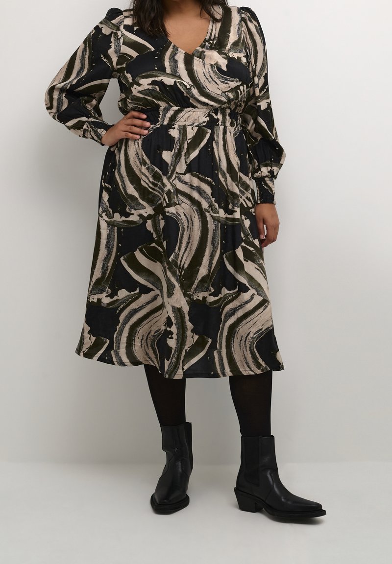Robe à manches longues avec un décolleté en V profond, présentant un motif abstrait tourbillonnant en noir, beige et vert olive, cintrée à la taille. Bottines noires à la cheville.