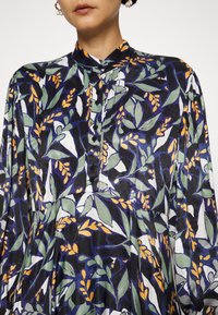 Femme portant une robe à col haut boutonnée avec des manches longues présentant un motif abstrait audacieux de feuilles bleu, vert, orange et blanc.