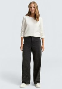 Zwarte wijde jeans met studs aan de voorkant, gecombineerd met een crème kleurig shirt met lange mouwen. Model staat tegen een lichtgrijze achtergrond.