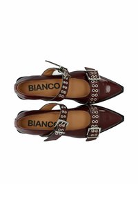 Sapatos de couro verniz bordô com biqueira apontada, apresentando fivelas prateadas e ilhós decorativos ao longo das correias. Logótipo da marca visível no interior.