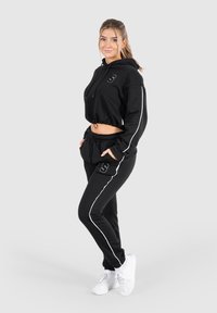 Schwarzer, verkürzter Hoodie mit weißen Akzenten und Kordelzug, kombiniert mit schwarzen Sweatpants, die einen weißen Seitenstreifen und ein Logo haben. Weiße Sportschuhe.