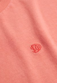 Koralfarvet kortærmet t-shirt lavet af blød bomuld, med et diskret broderet logo i midten. Glat tekstur og klassisk v-udskæring.