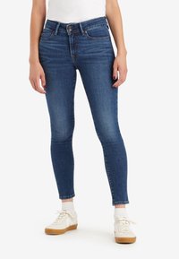 Jeans ajustados de mezclilla azul con una cintura de altura media, cinco bolsillos y un acabado desgastado; combinados con zapatillas blancas y calcetines de longitud media.