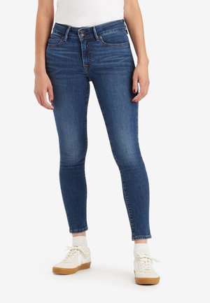 Jeans ajustados de mezclilla azul con una cintura de altura media, cinco bolsillos y un acabado desgastado; combinados con zapatillas blancas y calcetines de longitud media.