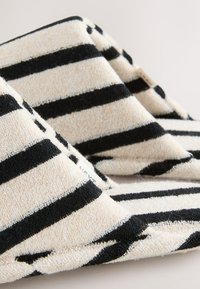 Gros plan sur des chaussons en tissu doux à rayures horizontales noires et blanches avec un design bas et ouvert à l'arrière sur un fond uni.