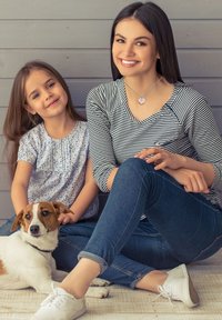 Kvinde i en stribet top og med halskæde sidder ved siden af en pige i en blomstret bluse, og begge smiler. En lille hund hviler på gulvet foran.