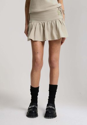 Person trägt beige Rüschen-Minirock, schwarze geraffte Socken und schwarze klobige Slip-On-Schuhe mit weißem Text auf den Riemen.