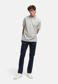 Grå polo skjorte med korte ermer, en spiss krage og et grønt logo-lapp, kombinert med marineblå bukser og hvite joggesko.