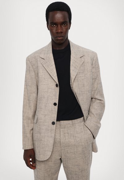 Lichtgrijze gestructureerde blazer met een geknipt revers en zwarte knopen, gedragen over een zwart shirt met korte mouwen. Getailleerde pasvorm, soepel materiaal.