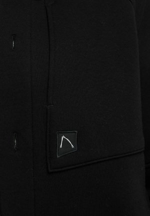 Schwarzer Pullover mit Knopfverschluss und einer aufgenähten Tasche mit minimalistischem weißem Logo. Hergestellt aus weichem, strukturiertem Stoff.