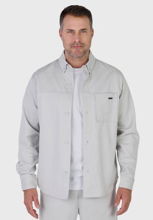 Mann mit kurzem Haar und Stoppelbart, trägt ein hellgraues Hemd über einem weißen T-Shirt vor einfarbigem Hintergrund.