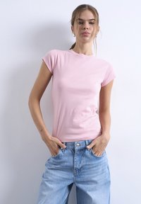 Νεαρή γυναίκα που στέκεται δίπλα σε τοίχο, φοράει ροζ ανοιχτόχρωμο κοντομάνικο πουκάμισο και φαρδιά μπλε τζιν, με τα χέρια στις τσέπες, κοιτάζοντας την κάμερα.