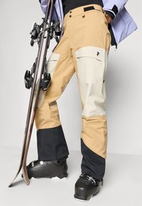 Beige en crème skibroek met zwarte accenten, uitgevoerd in waterdicht materiaal, functionele zakken en een losse pasvorm. Gecombineerd met zwarte skischoenen.