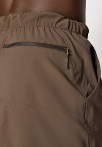 Nærbillede af brune elastiske bukser med sidelommer og lynlåslommer bagpå på en mørk hudbaggrund.