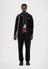 Veste zippée en velours noir avec des accents blancs, t-shirt graphique noir, pantalons noirs et chaussures noires. Sac épaule gris avec bandoulière.