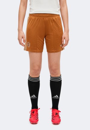 Un modèle porte un short de football marron, des chaussettes Adidas noires montant jusqu’aux genoux et des chaussures de football rouge vif, et se tient devant un fond uni.