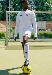 Jersey de deporte de manga larga blanca con un patrón geométrico texturizado, pantalones cortos blancos, botas de fútbol negras y de pie sobre un balón de fútbol amarillo.