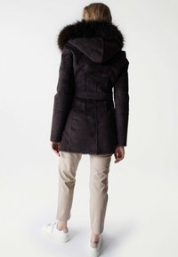 Persona de pie con la espalda visible, vestida con un abrigo con capucha de color marrón oscuro con borde de piel, pantalones beige y zapatillas blancas sobre un fondo blanco.